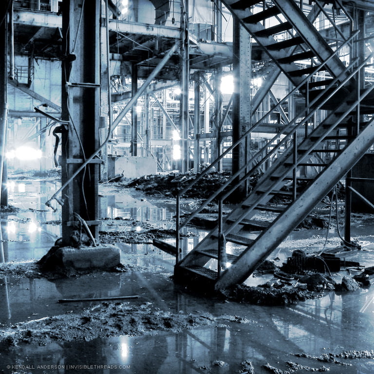 Metal stairs rise from the ground floor to the second level of an abandoned industrial power station. The ground is muddy and covered with pools of water. Exposed steel columns and beams recede into the distance.