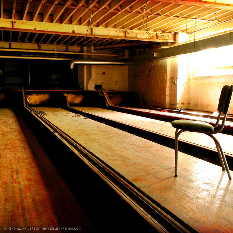 A single chair is placed in the middle of a bowling alley lane. The bowling alley is in the basement of a building, and an orange glow covers everything from the sunlight coming through the basement windows. The bowling lanes are dirty and covered in dust.