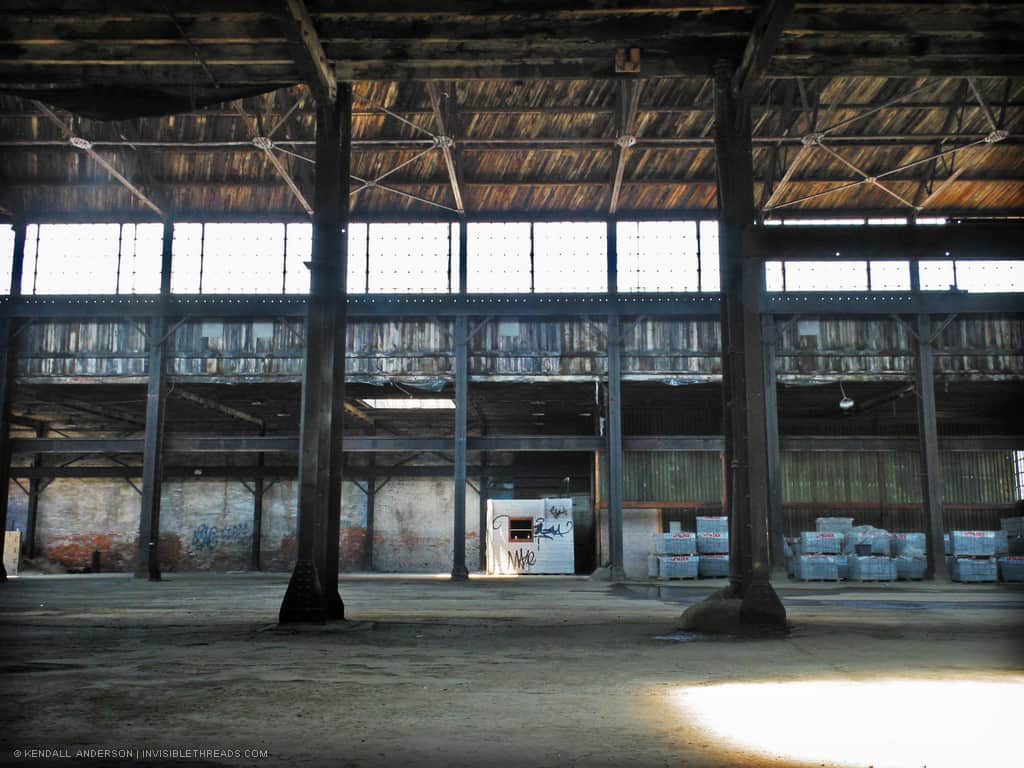 Industrial warehouse interior, with steel columns supporting trusses and a wood roof, illuminated by skylights