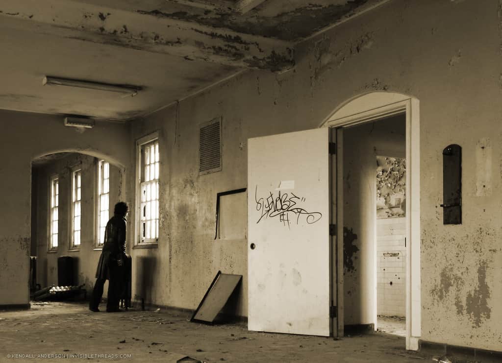 Inside an abandoned room with graffiti and peeling paint, a woman peers out one of the windows to look outside.