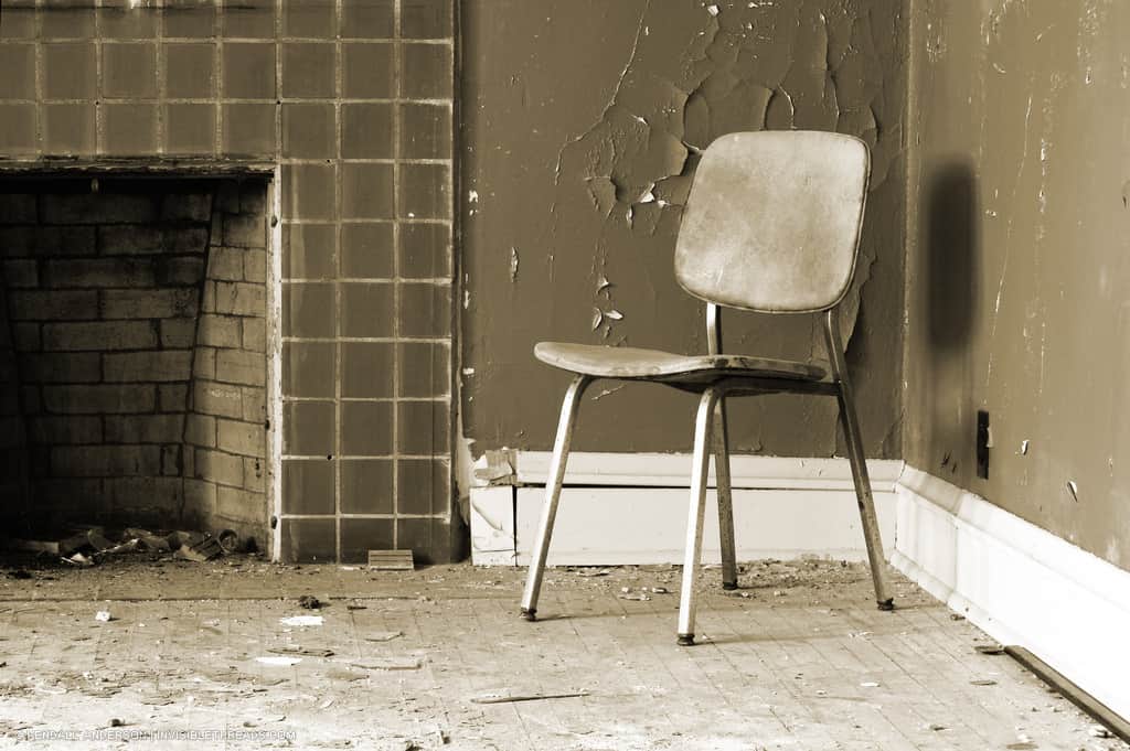 A single chair sits in a corner beside a fireplace. The paint on the walls is beginning to crack and peel.