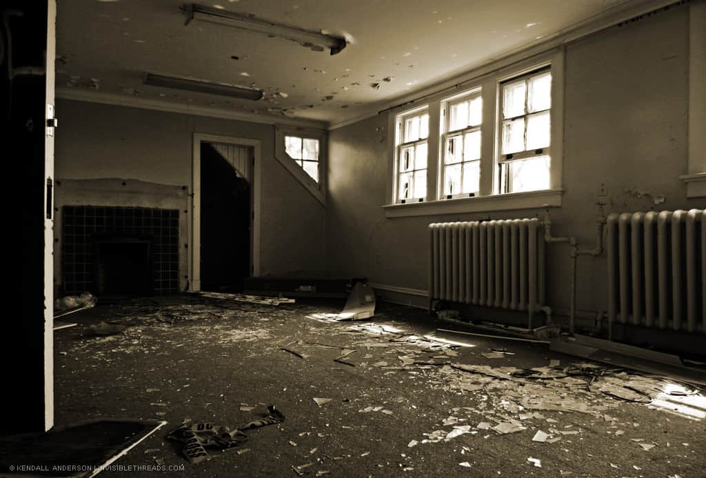 An interior room has metal radiators on the walls under the window. Broken glass and plaster are strewn about the floor. An empty fireplace is on one wall.