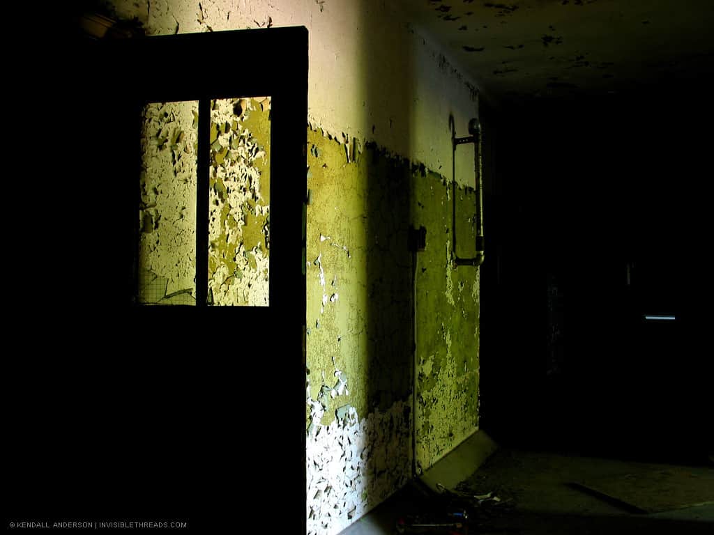 The silhouette of an open door with windows is seen against a green and white wall. The wall is crumbling and covered in peeling, cracking paint.