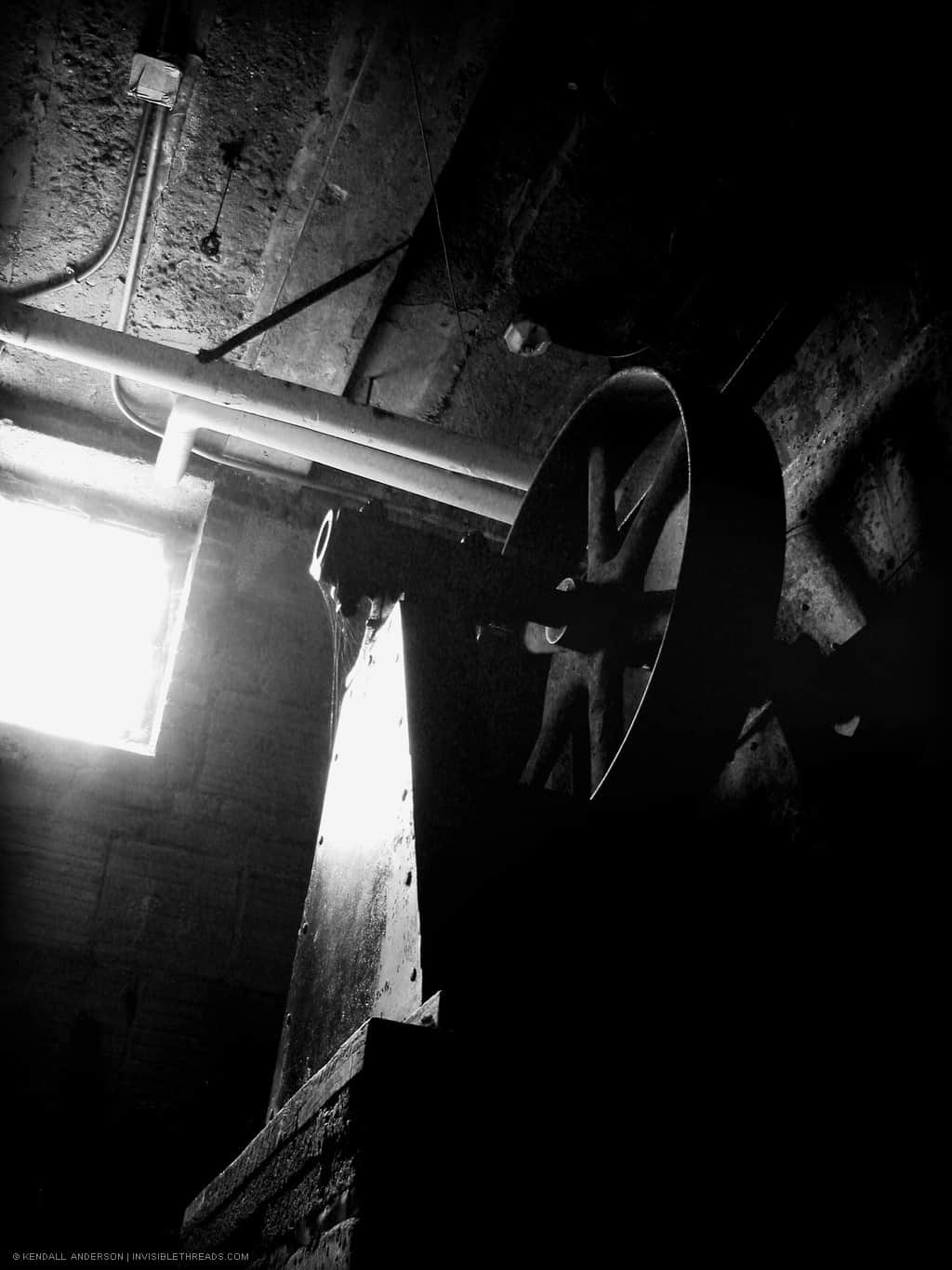 A large metal wheel is attached to some mechanical equipment in a dark basement room.