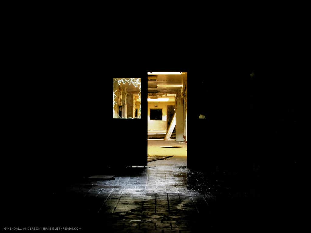 The wet reflection of a tile floor is visible in an otherwise pitch black room. Through a door opening and broken glass, the bright yellow corridor beyond can be seen.