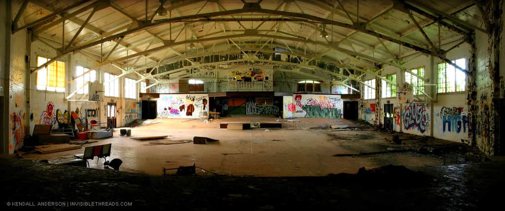 The interior of a large gymnasium space has walls covered with graffiti. Debris is on the floor.