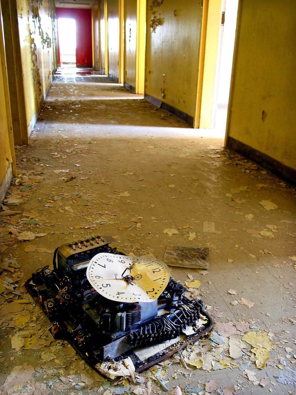 A long yellow hallway allows light to shine on to the floor. A large wall-mounted clock lies in the middle of the floor, broken. The floor is covered in debris and paint chips.