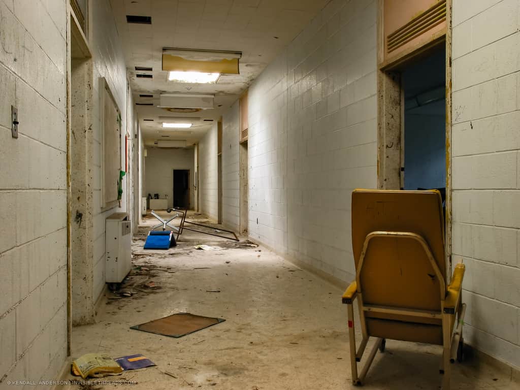 An empty corridor lined with concrete block walls is filled with debris and overturned chairs. A large chair is in the hallway facing an open entrance to a room.