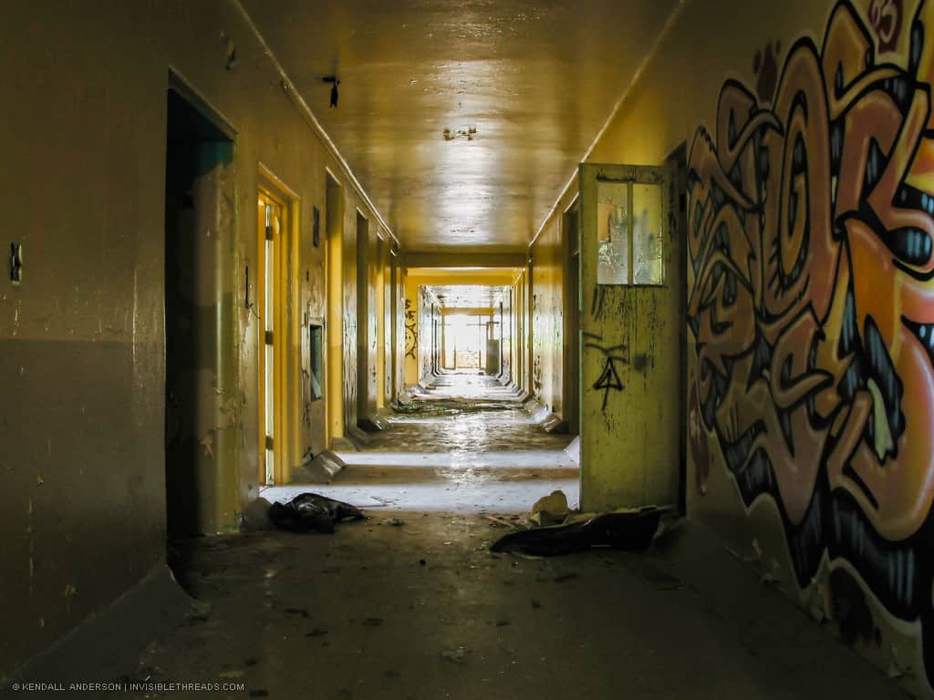 A dark yellow corridor has a large piece of graffiti on the wall. Patterns of light and dark illuminate the hallway from the rooms to each side. Debris is strewn about the corridor.