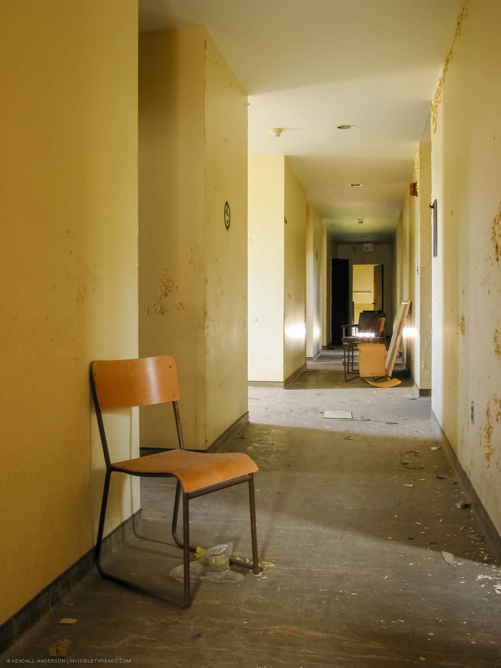 A chair is on the floor in a long yellow hallway, with additional chairs piled in the distance. The floor has debris on it and the paint on the walls is just beginning to show signs of water damage and peeling.
