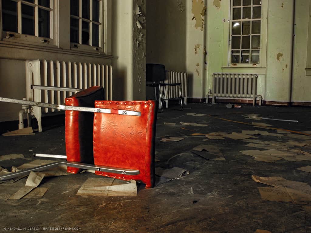 A single orange chair is tipped over inside a room with boarded up windows. Old papers are strewn about the floor, and the paint is peeling off the walls.