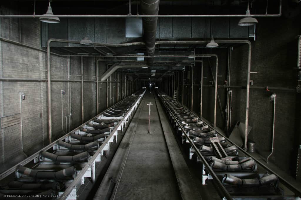A dark square tunnel recedes into the distance, into complete darkness. There is room to walk down the middle, and on both the left and right sides are conveyor belts stretching into the darkness. Ductwork and lamps hang from the ceiling.