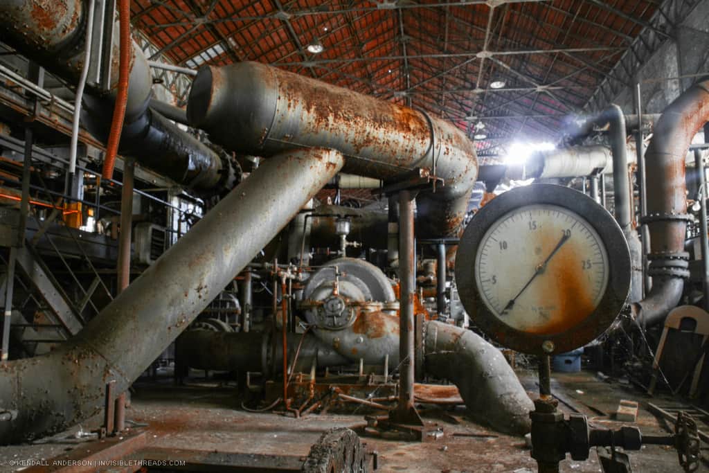 A dense array of metal pipes of varying sizes connect into each other and twist around the interior of an industrial building. A large pressure gauge is in the foreground with a reading of 0.