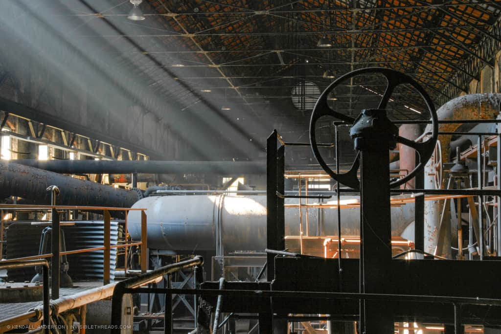 Shafts of light shine into the interior of a dark industrial building. The light reveals large metal pipes and railings densely packed into the building. The silhouette of a large valve wheel is in the foreground.