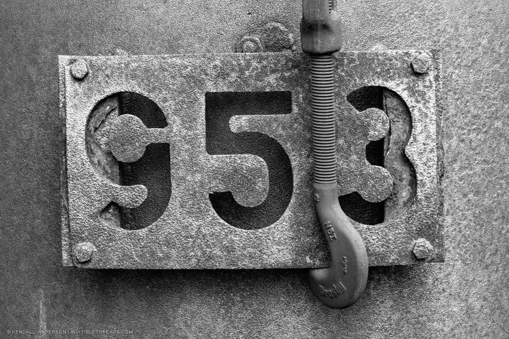 A rusted metal plate is in front of a textured metal surface. The plate has the number 953 cut out of the surface.