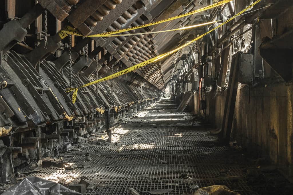 A derelict tunnel lined with steel metal grating runs at the bottom of an industrial coke oven. The tunnel has a triangular cross section, and yellow caution tape blocks passage.