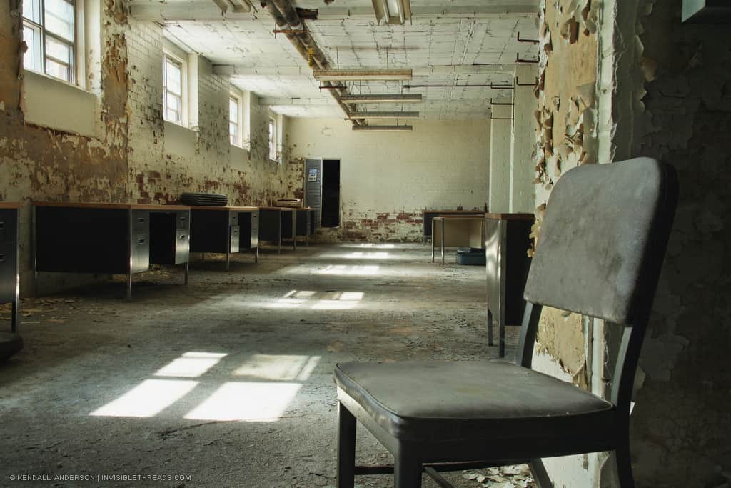 A single office chair sits at the entrance of an abandoned basement office room with natural sunlight from windows. Walls, columns and roof have peeling paint, and office desks are lined up against the wall.