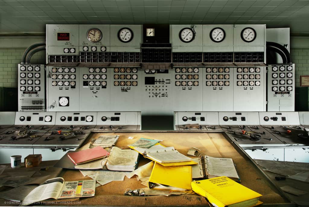 The gauges and instruments in an industrial control room are mounted on a large floor to ceiling panel. Operator's controls are in front of the panel. A supervisor's desk oversees the entire room, with multiple binders strewn about the desk, some open. The room is dirty and abandoned.