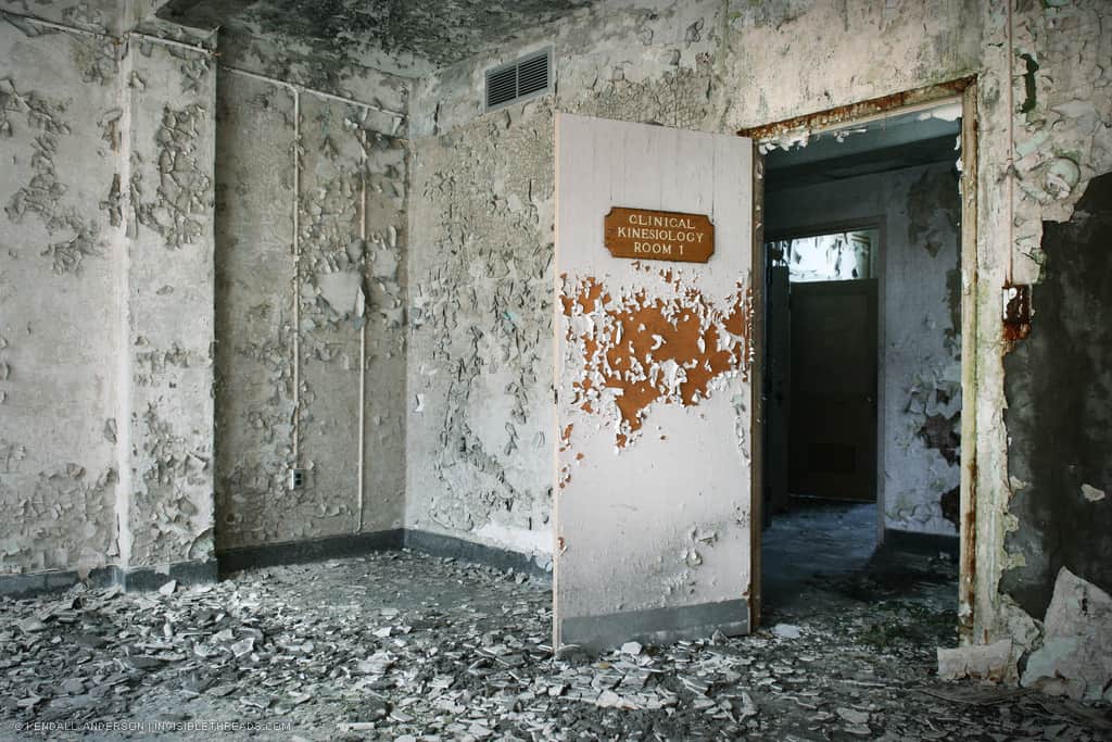 An open door shows the exit from a derelict room with debris. White paint on the walls and doors is peeling everywhere, revealing orange paint beneath.