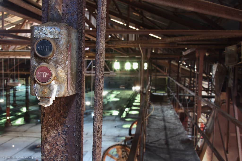 Rusting receptacle box with push buttons labelled 'Start' and 'Stop', beside a catwalk in an abandoned brickmaking factory
