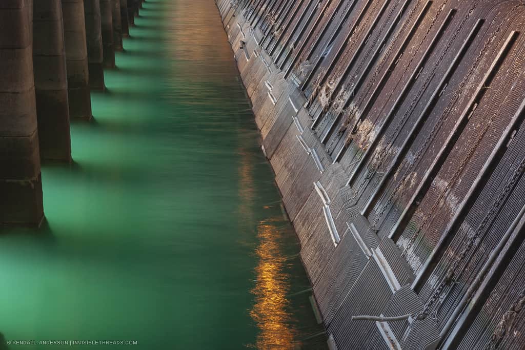 A channel of green water is adjacent to a steel surface protruding from the water at a 45 degree angle. This steel surface is a series of metal grates that prevent ice, trash and debris from entering the penstocks below.