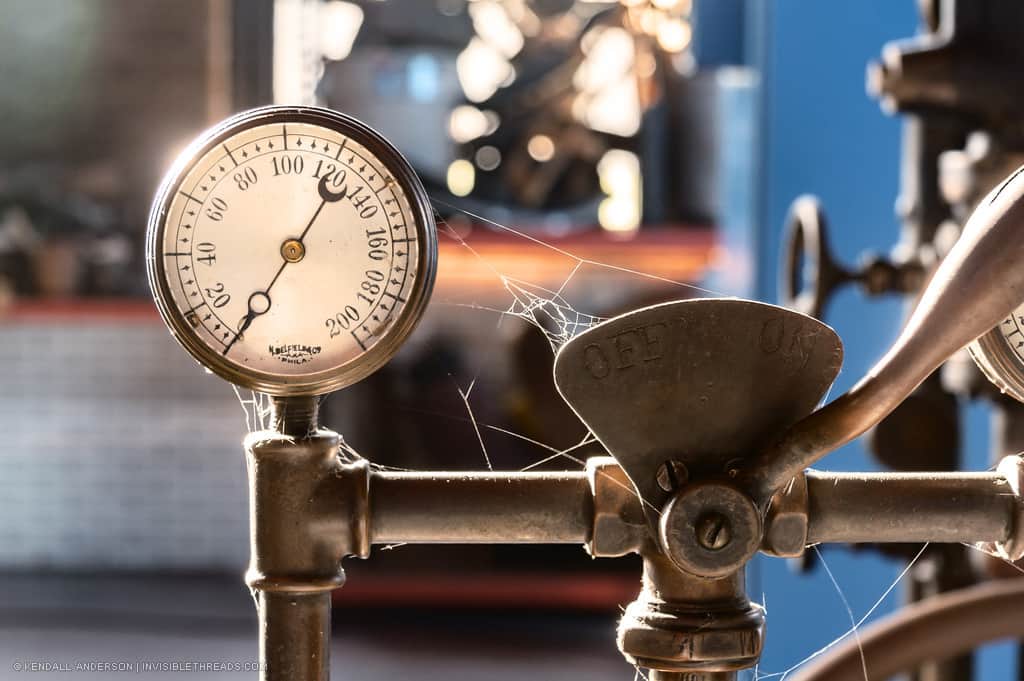 A gauge connected to some pipes is in direct sunlight, which is gleaming off the edges. Spiderwebs span from the gauge to the pipes. The gauge has a measurement reading of 0.