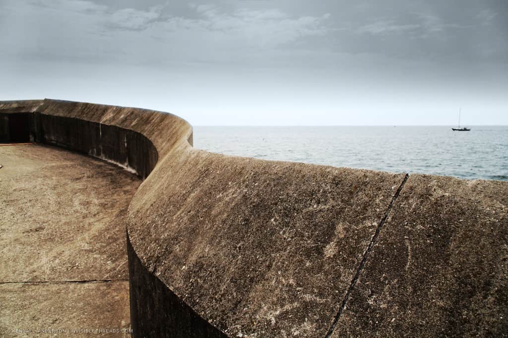 A curved concrete pier and wall, with Lake Ontario's water behind. A small sailboat is far off on the distance on the lake.