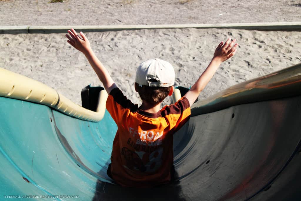 A small boy wearing a baseball hat and orange shirt is sliding down a metal slide in a playground, towards the sandy ground.
