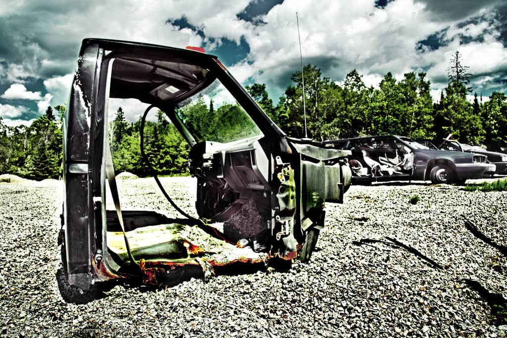 A stripped down metal cab from a pickup truck sits on a bed of gravel by itself. The doors and undercarriage have been removed, along with the interior seats. Junked cars are in the background.