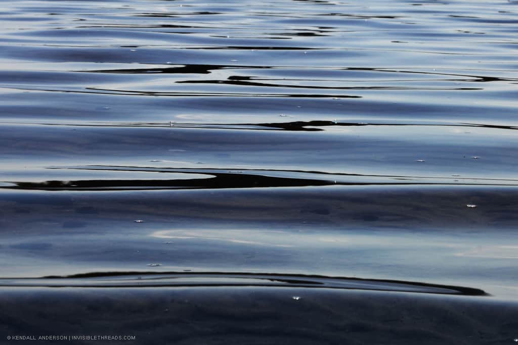 Blue calm lake water with white and black reflections, creating a linear pattern.