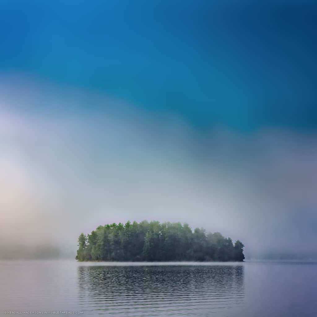 A single island covered with trees is surrounded by fog and mist on the lake water. The sky is also smooth with fog. The reflection of the island and trees is in the water in front of the island.