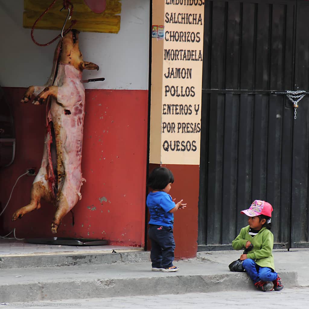 Two children play on the sidewalk outside a butcher shop, where a pig carcass hangs on the wall.