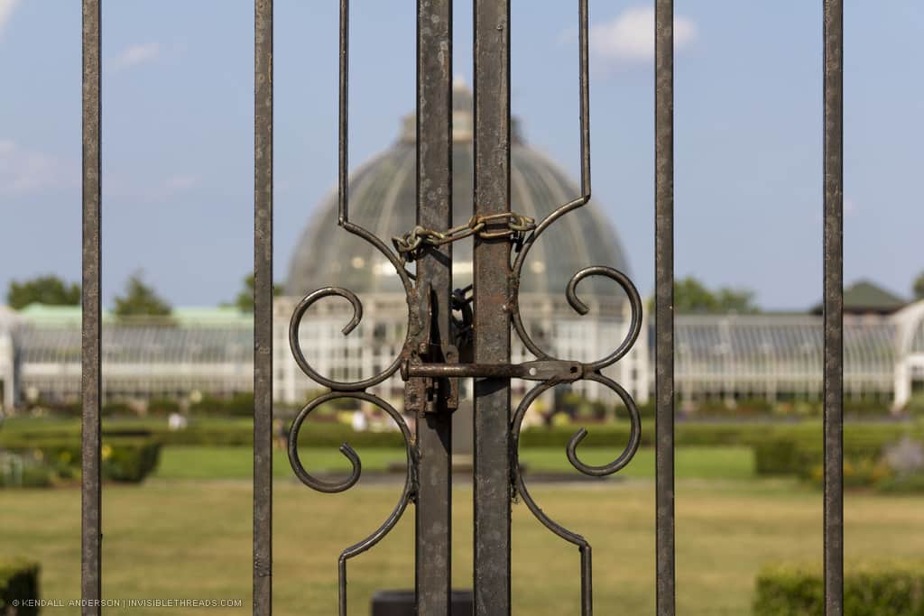 Close-up of a fence gate, locked with a chain, with a blurry conservatory building beyond