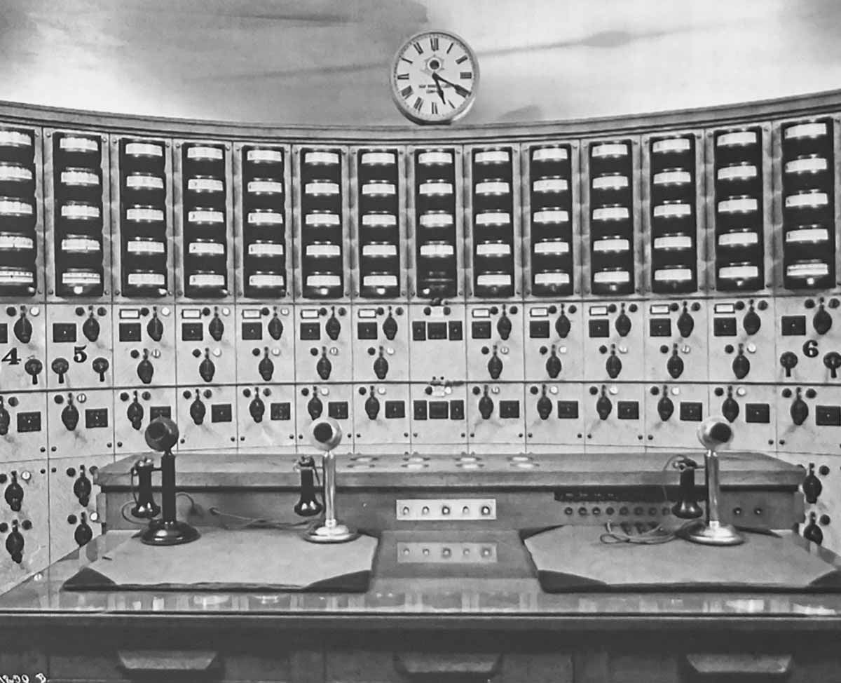 A power station control panel is covered with meters, gauges and switches all arranged in columns (one column per alternator). A wooden desk with microphones is in front of the control panel. Archival photo from some time near 1910.
