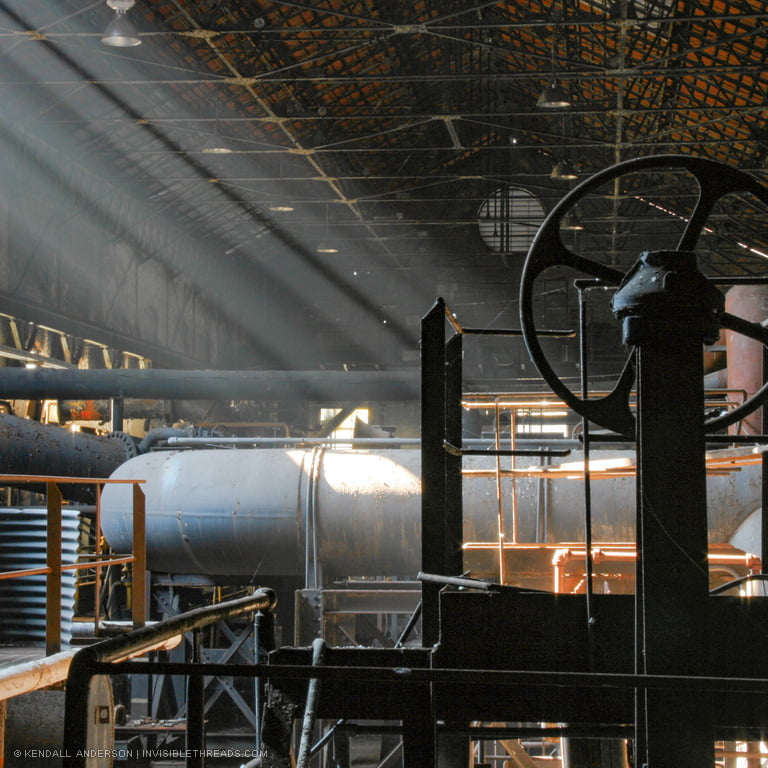 Shafts of light shine into the interior of a dark industrial building. The light reveals large metal pipes and railings densely packed into the building. The silhouette of a large valve wheel is in the foreground.
