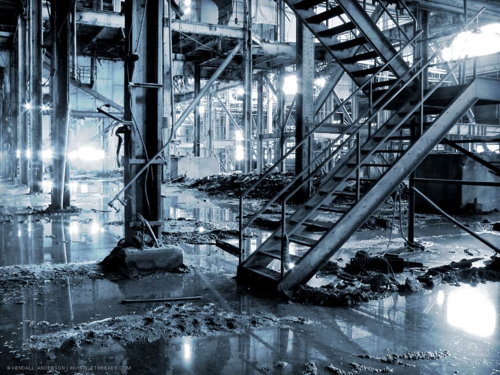Metal stairs rise from the ground floor to the second level of an abandoned industrial power station. The ground is muddy and covered with pools of water. Exposed steel columns and beams recede into the distance.