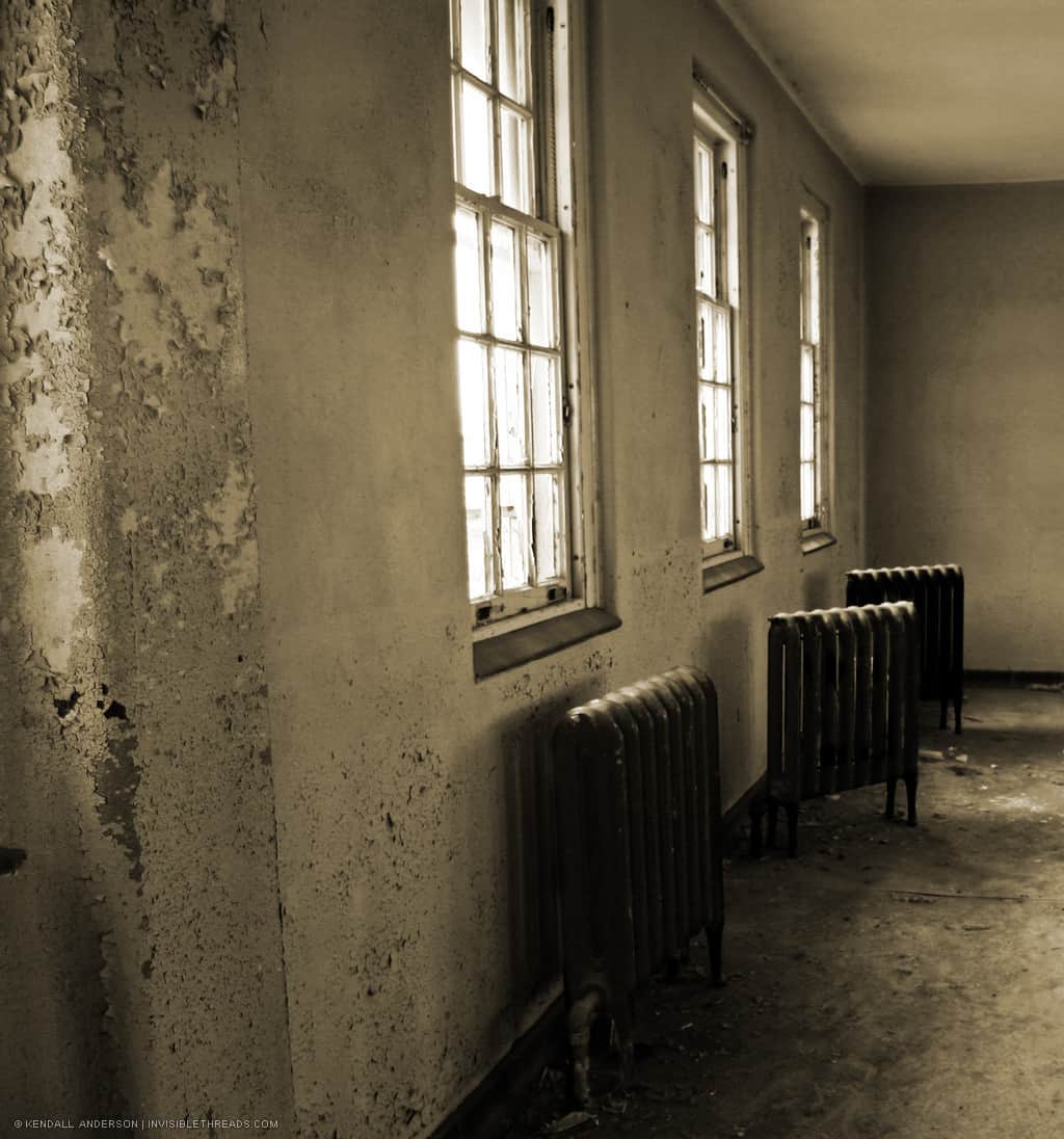 Three metal radiators rest against the wall under three windows, in an abandoned room where the paint is peeling off the walls.