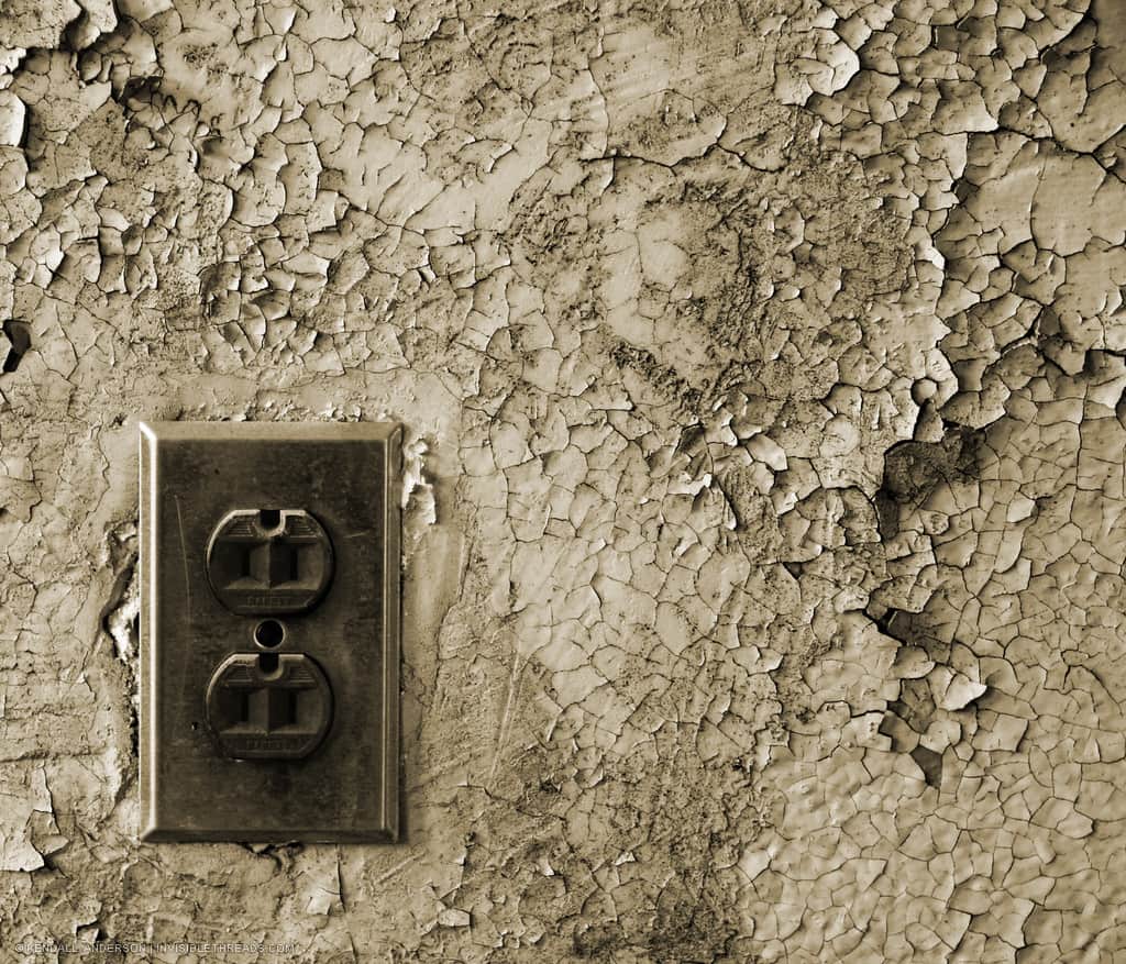An electric outlet receptacle is on a wall completely covered with cracked and peeling paint.