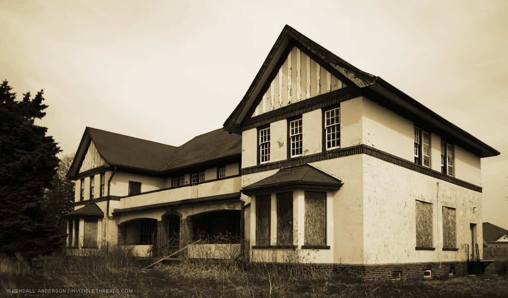A 2-storey institutional cottage building is surrounded by overgrown grass. All windows on the ground floor are boarded up.