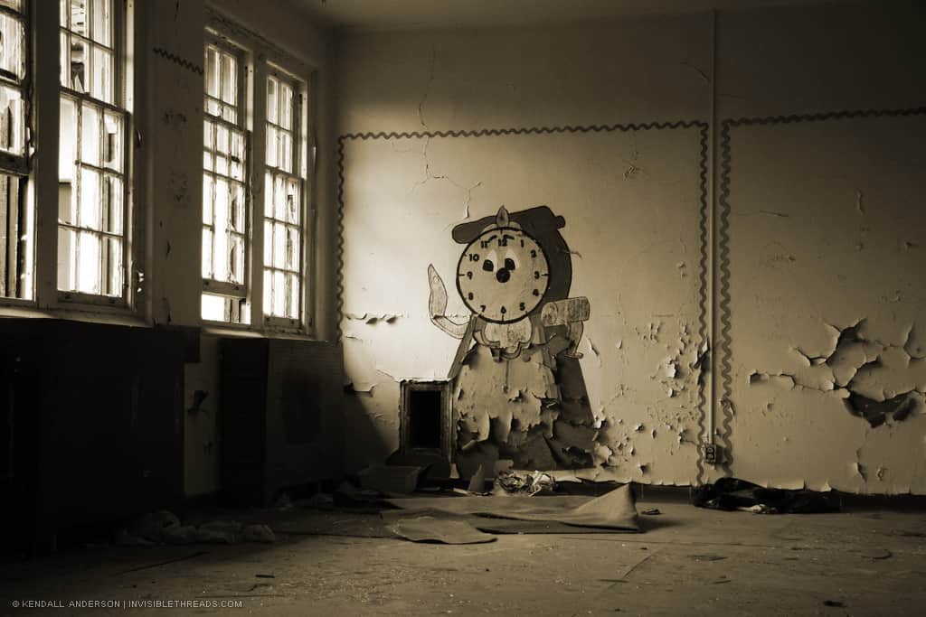 Inside an abandoned room, a character resembling a clock is painted on the wall. Windows allow some light into the room and highlight the peeling paint on the walls.