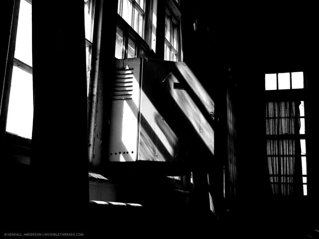 Shadows are visible against a window mounted air conditioner inside a dark room with windows and curtains.
