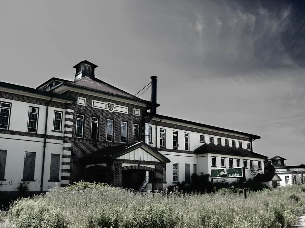 The front facade of a 2-storey institutional building is surrounding by overgrown vegetation. Most windows on the ground floor are boarded up.