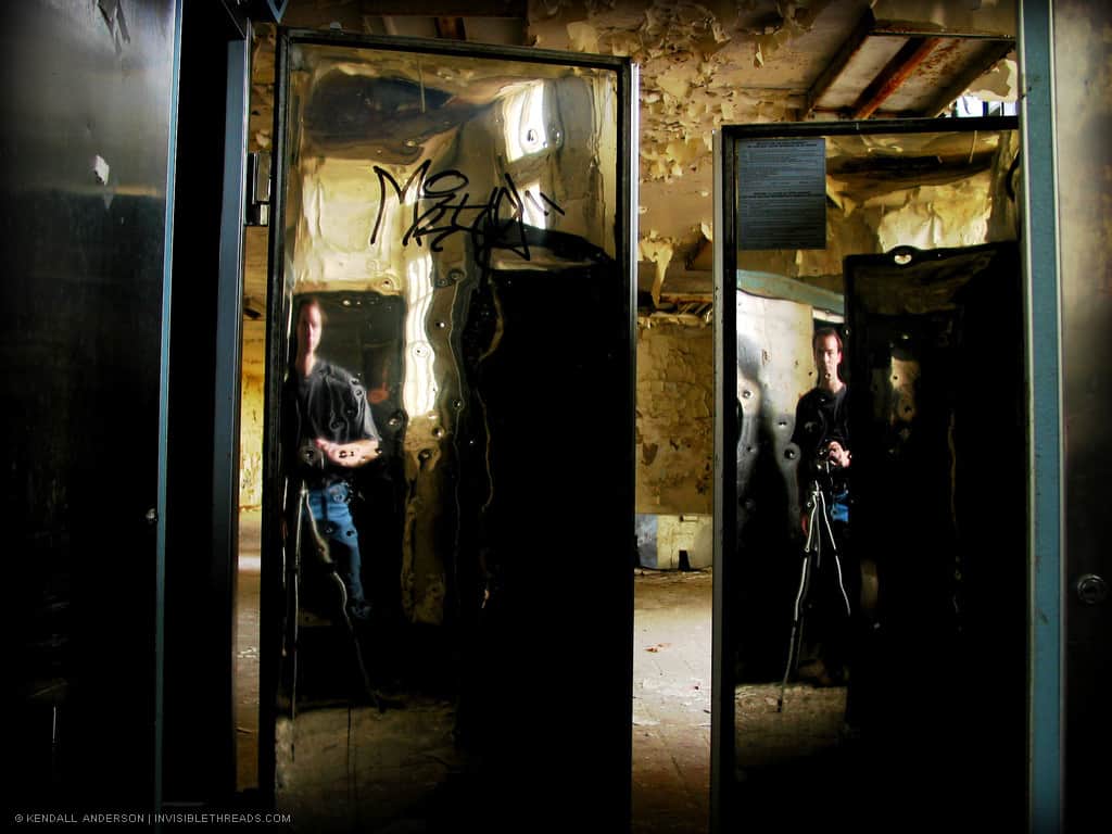 The reflection of a person is seen in the rippled stainless steel doors of refridgeration units inside an abandoned room. The ceiling and walls behind are covered in peeling paint.