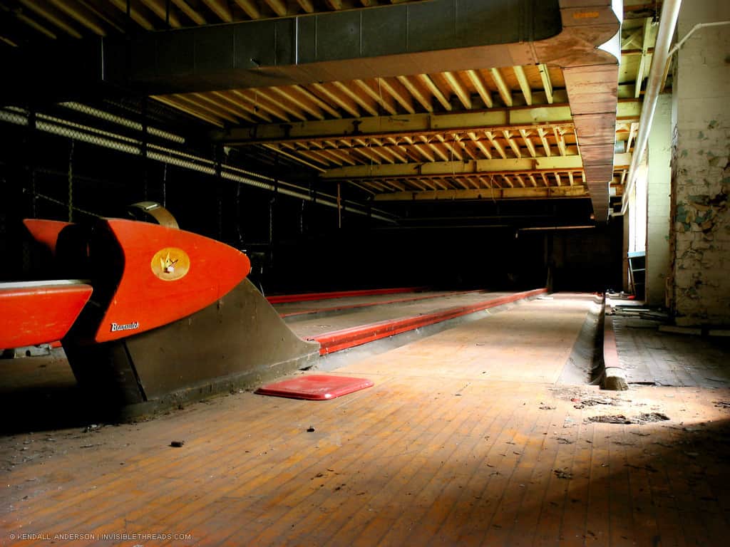 The red ball return equipment is seen at the start of a bowling alley in the basement of a recreation centre building. The space is lit by the windows at ground level.