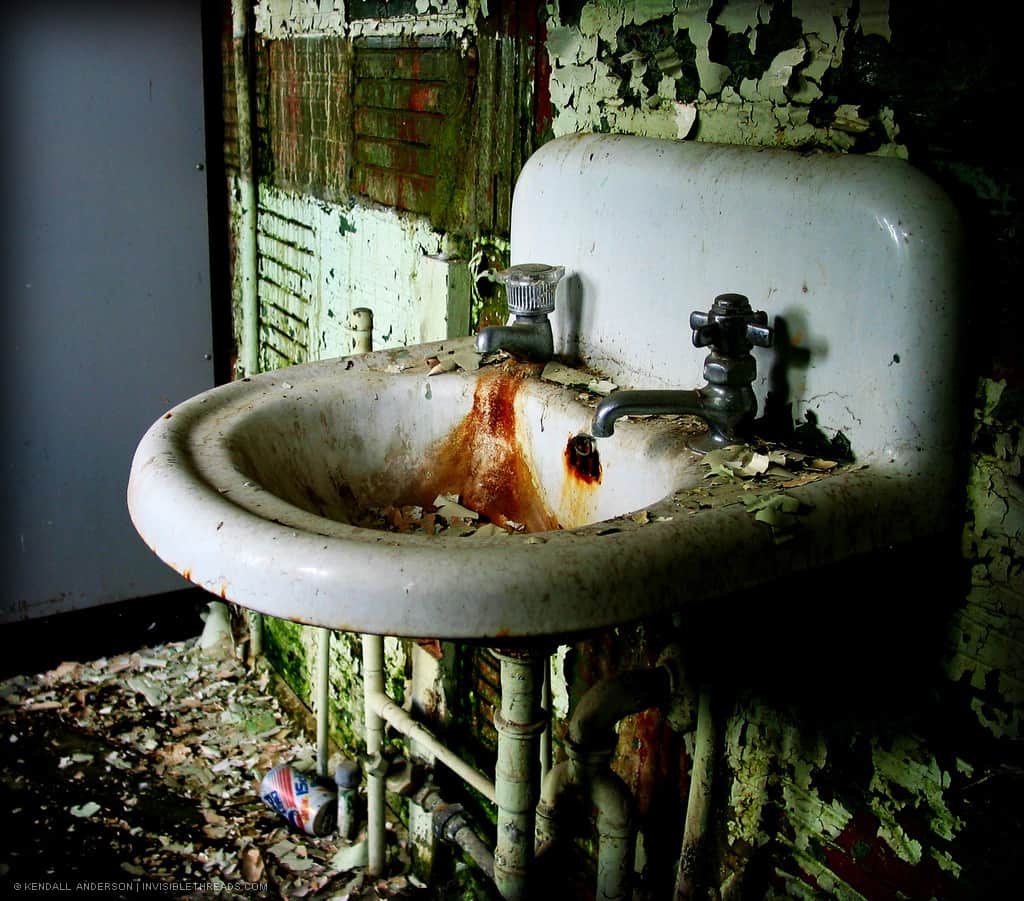 A white ceramic sink has rust stains and is filled with flakes of paint. The wall behind is dirty, grimy and full of decay and debris.