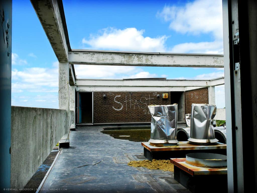 The cloud filled blue sky is seen from the rooftop. Concrete beams are above, and a brick wall contains two doors leading down inside the building.