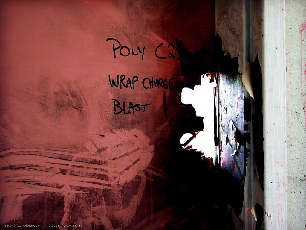A red door with bootprints looks like the lock has been blasted off. Someone has written on the door in marker, reading 'Poly C2 Wrap Charge Blast'.