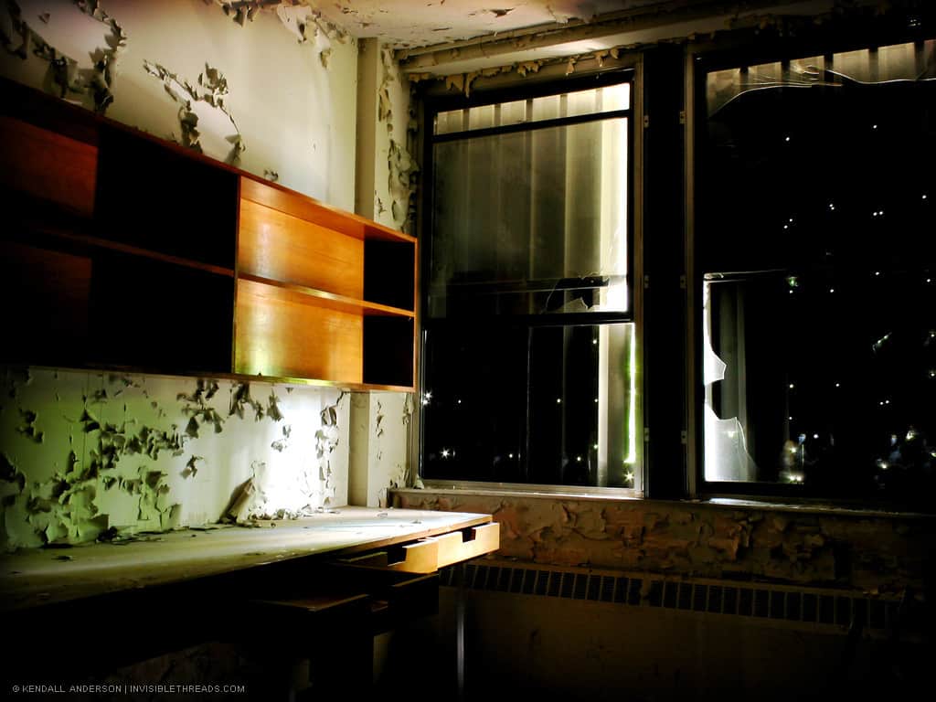 The interior of a dark room contains a desk and cupboards. Some small points of light appear through the holes in the boarded up windows.