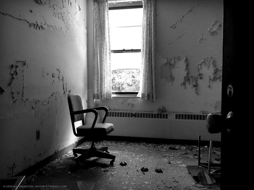 The interior of a decaying room with peeling paint and debris, has a single office chair sitting near the corner window.