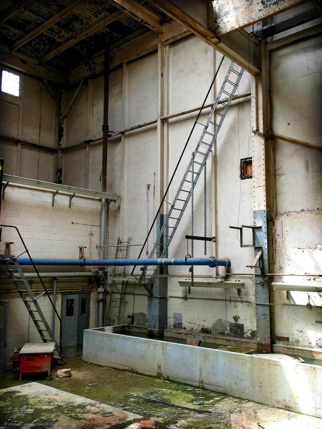 A steep narrow ladder extends from the ground floor up 3 storeys into the attic, with bars instead of stairs, and a minimal railing, inside a mechanical room.